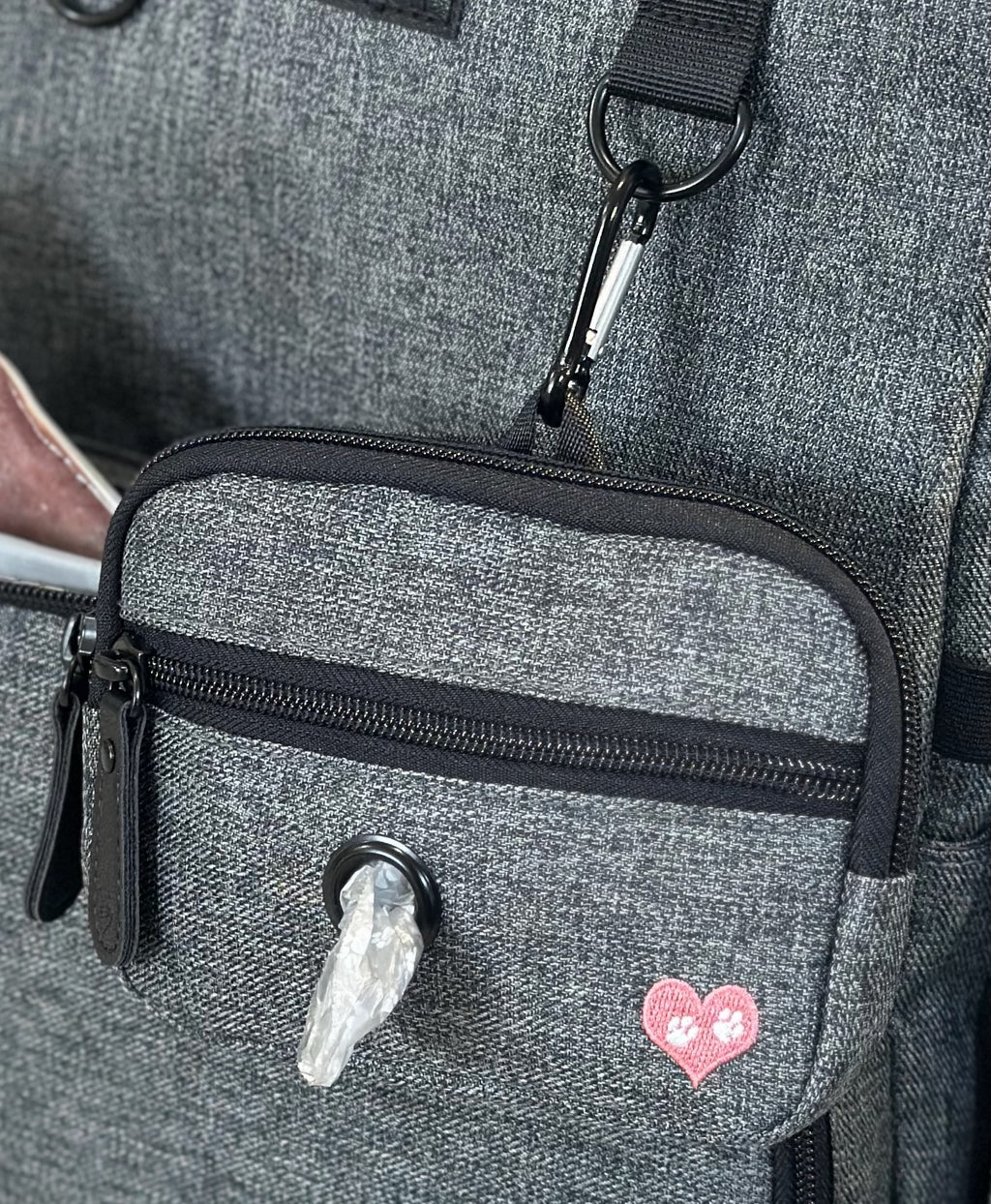 Close-up of a gray heathered fabric bag with a front zip pocket, black zipper trim, a black carabiner clip, and a small pink heart patch on the pocket.