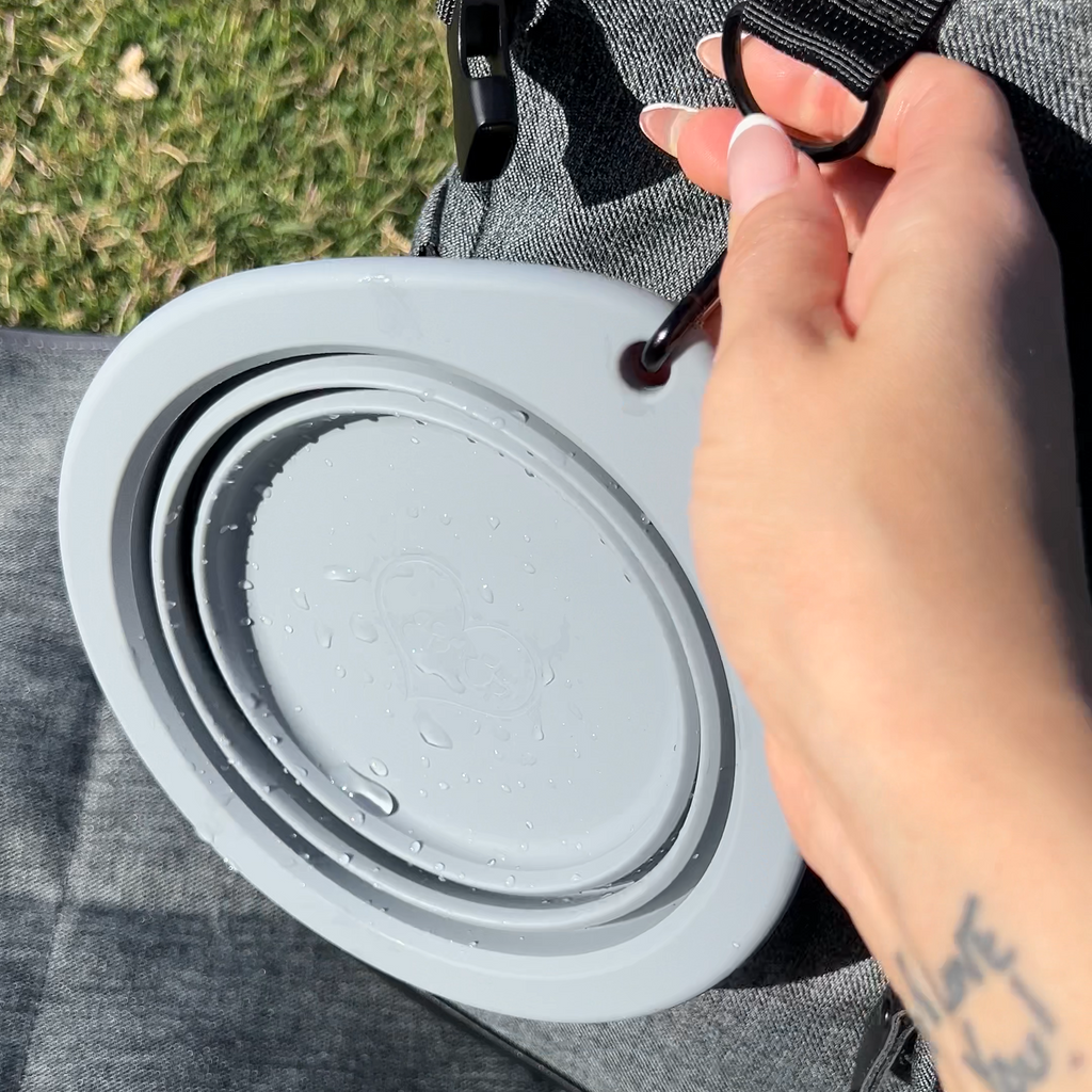 A close-up of a person's hand attaching a black strap to a gray collapsible travel water bottle container, placed on a gray fabric surface with grass in the background.